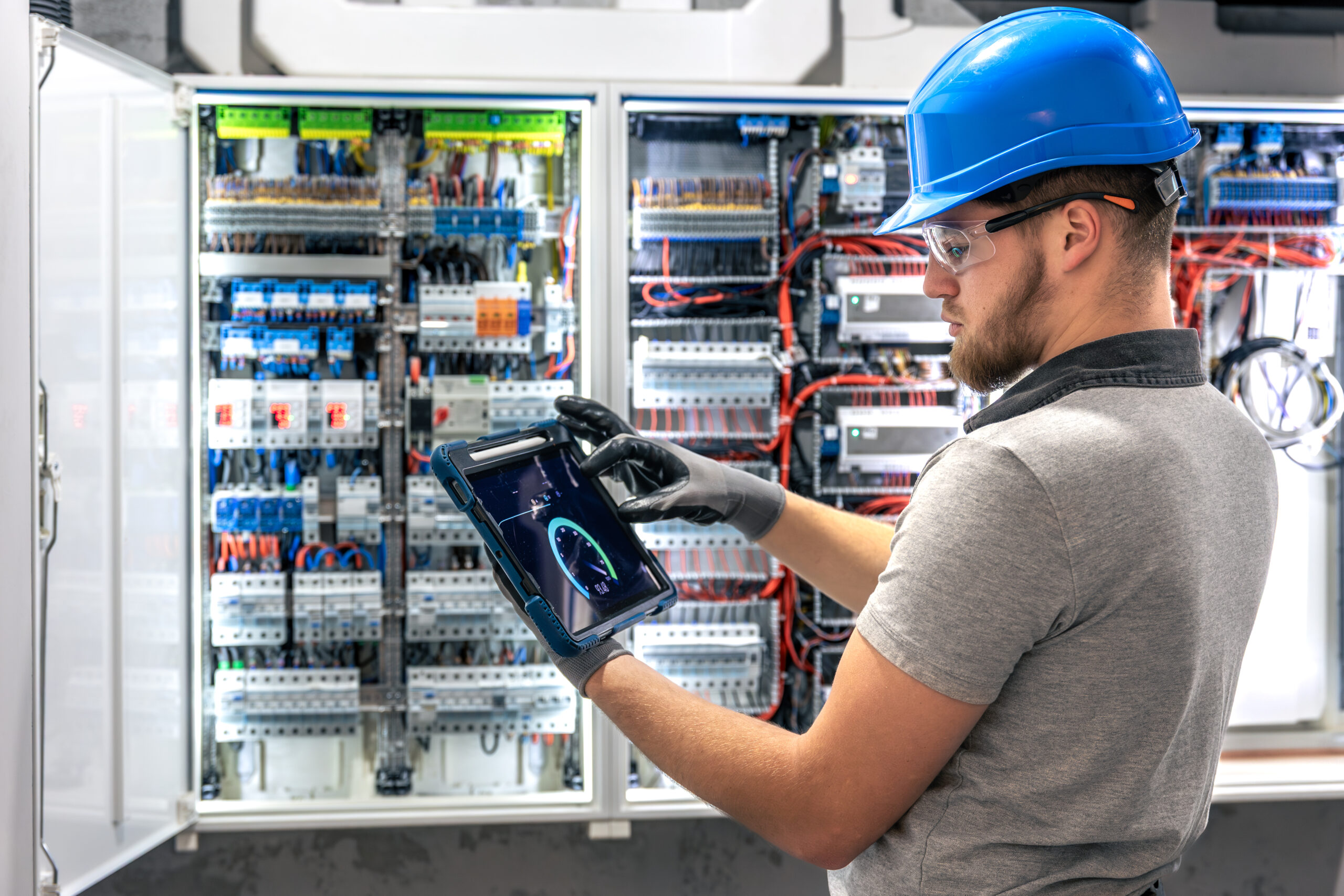 A man in protective gear holds a tablet and looks at the camera while standing in front of a power switchboard. A scene of modern technical work in an industrial environment.
